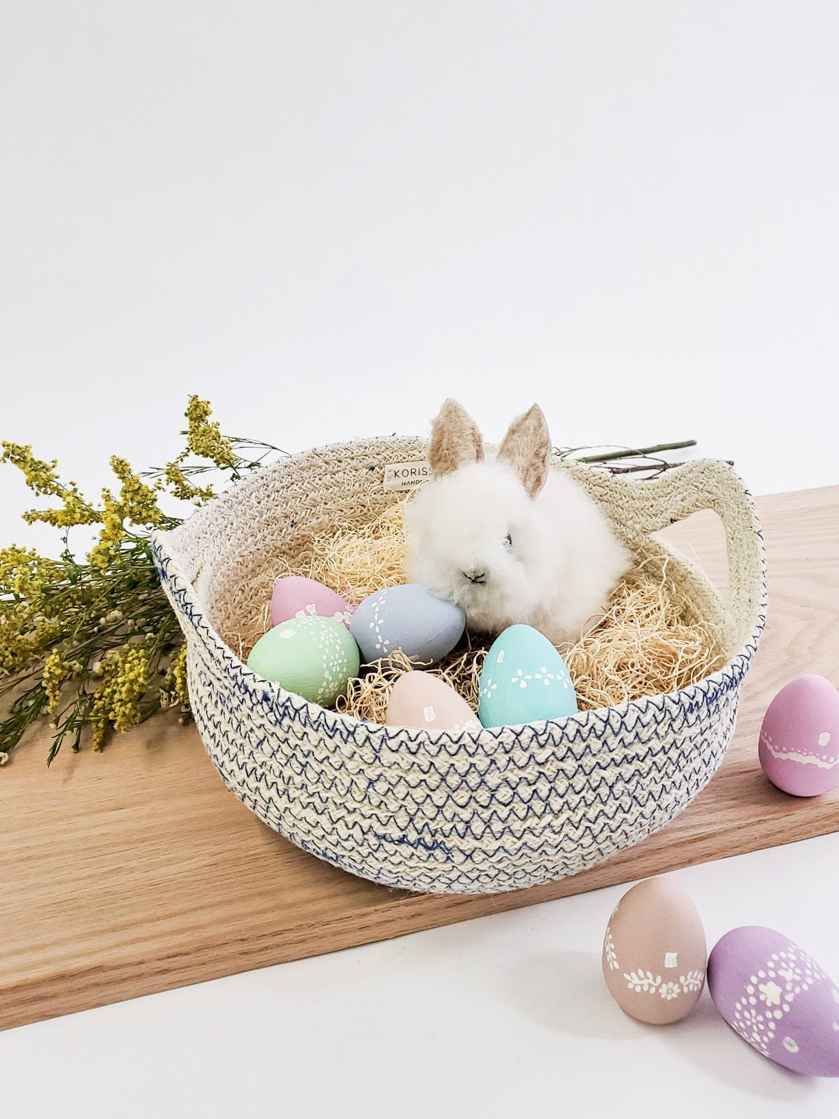 ALARA LOFTHandwoven off-white jute basket with blue stitching, filled with straw, pastel Easter eggs, and a fluffy bunny.