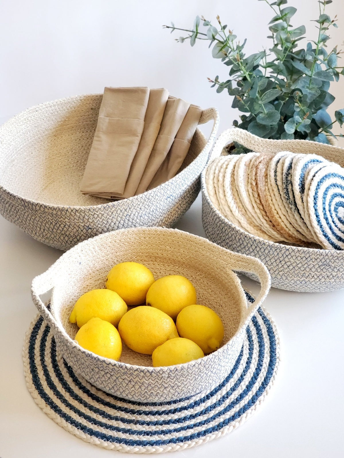 ALARA LOFTHandwoven jute fruit bowl with blue stitching, off-white color, featuring handles and a blue striped mat.